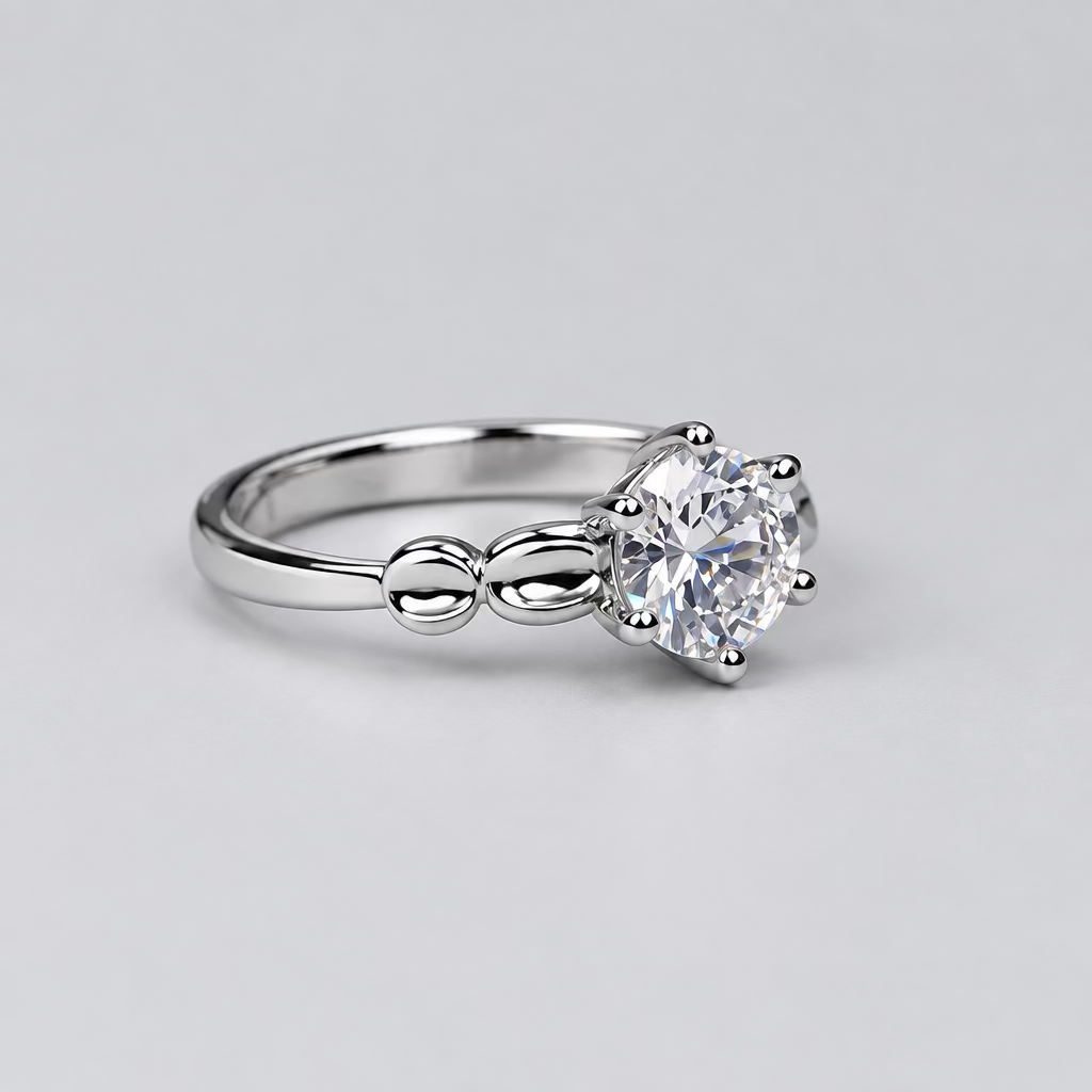 A silver-toned solitaire ring with a clear round gemstone, displayed on a light surface with a blurred white jewelry box featuring an 'M' logo in the background.