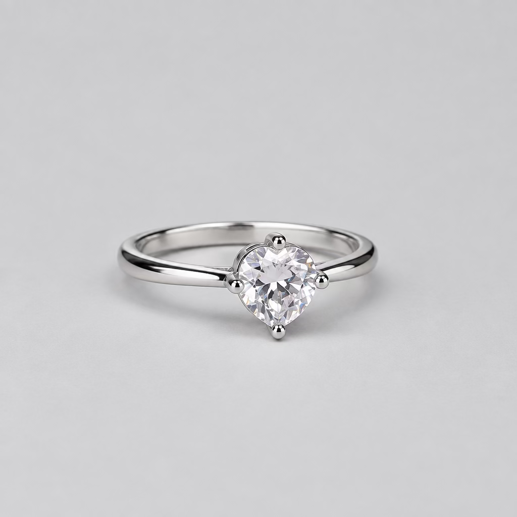 A silver solitaire engagement ring with a round-cut clear gemstone, displayed on a light lavender surface with a blurred white jewelry box featuring an orange 'M' logo in the background.