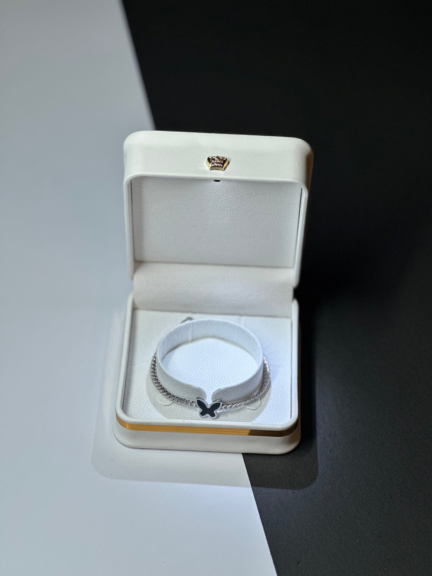 A silver-toned chain bracelet with a small black bow charm, displayed inside an open cream-colored jewelry box on a split white and black background.