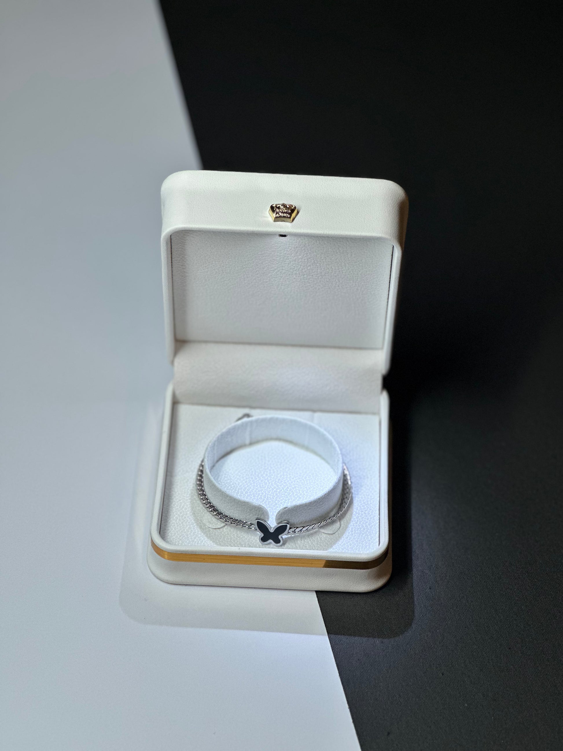 A silver-toned chain bracelet with a small black bow charm, displayed inside an open cream-colored jewelry box on a split white and black background.