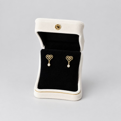 A pair of gold heart-shaped dangle earrings displayed in an open white jewelry box with a black velvet interior, set against a split black and white background.