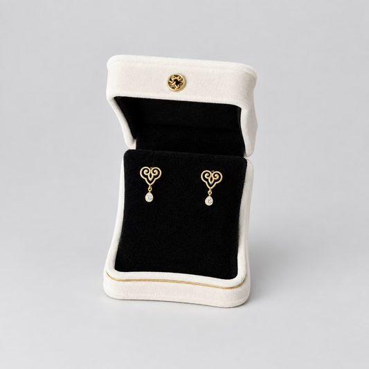 A pair of gold heart-shaped dangle earrings displayed in an open white jewelry box with a black velvet interior, set against a split black and white background.