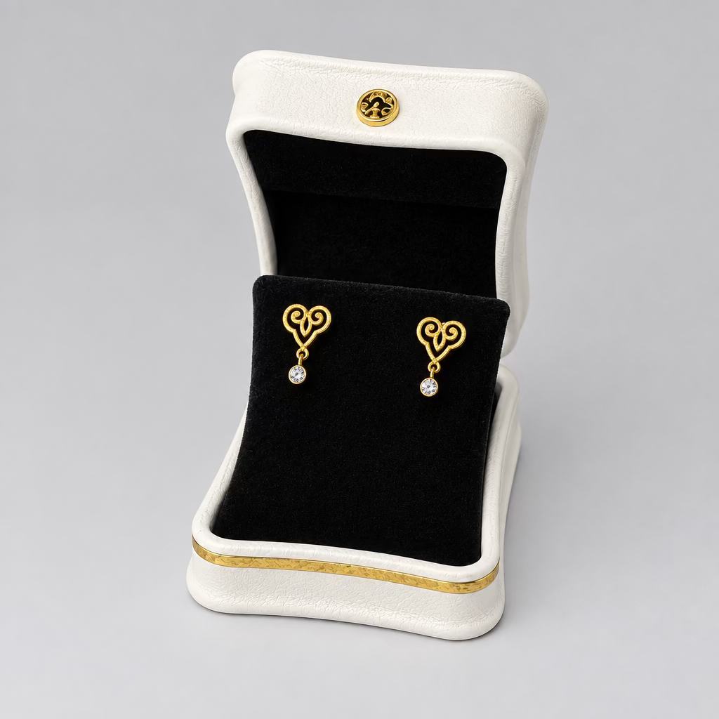 A pair of gold heart-shaped stud earrings with small clear stones, displayed in an open white jewelry box with a black velvet interior on a split black and white background.