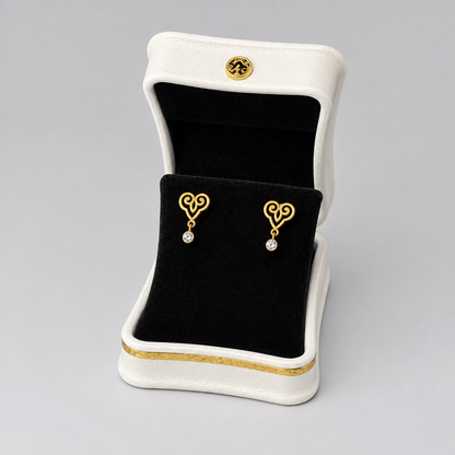 A pair of gold heart-shaped stud earrings with small clear stones, displayed in an open white jewelry box with a black velvet interior on a split black and white background.