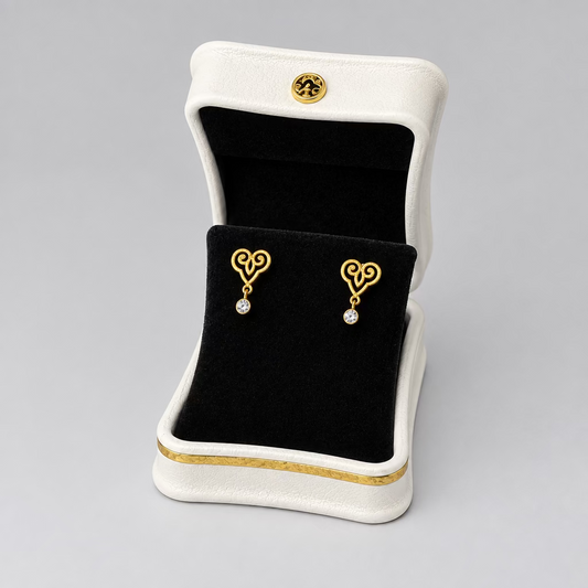 A pair of gold heart-shaped stud earrings with small clear stones, displayed in an open white jewelry box with a black velvet interior on a split black and white background.
