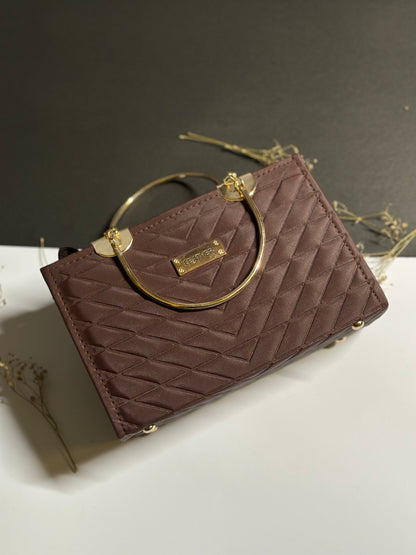 A small quilted handbag in a deep plum-brown color with a gold-tone circular top handle and a gold brand plaque. The bag is styled on a white surface with dried floral elements against a dark gray background.
