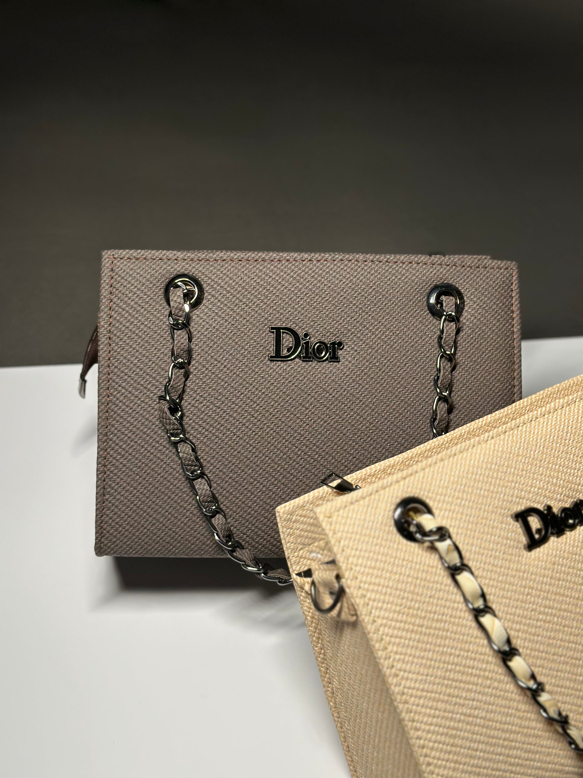 A Dior handbag in taupe textured leather with a silver-tone chain strap, shown alongside another beige Dior bag on a white surface against a dark gray background.