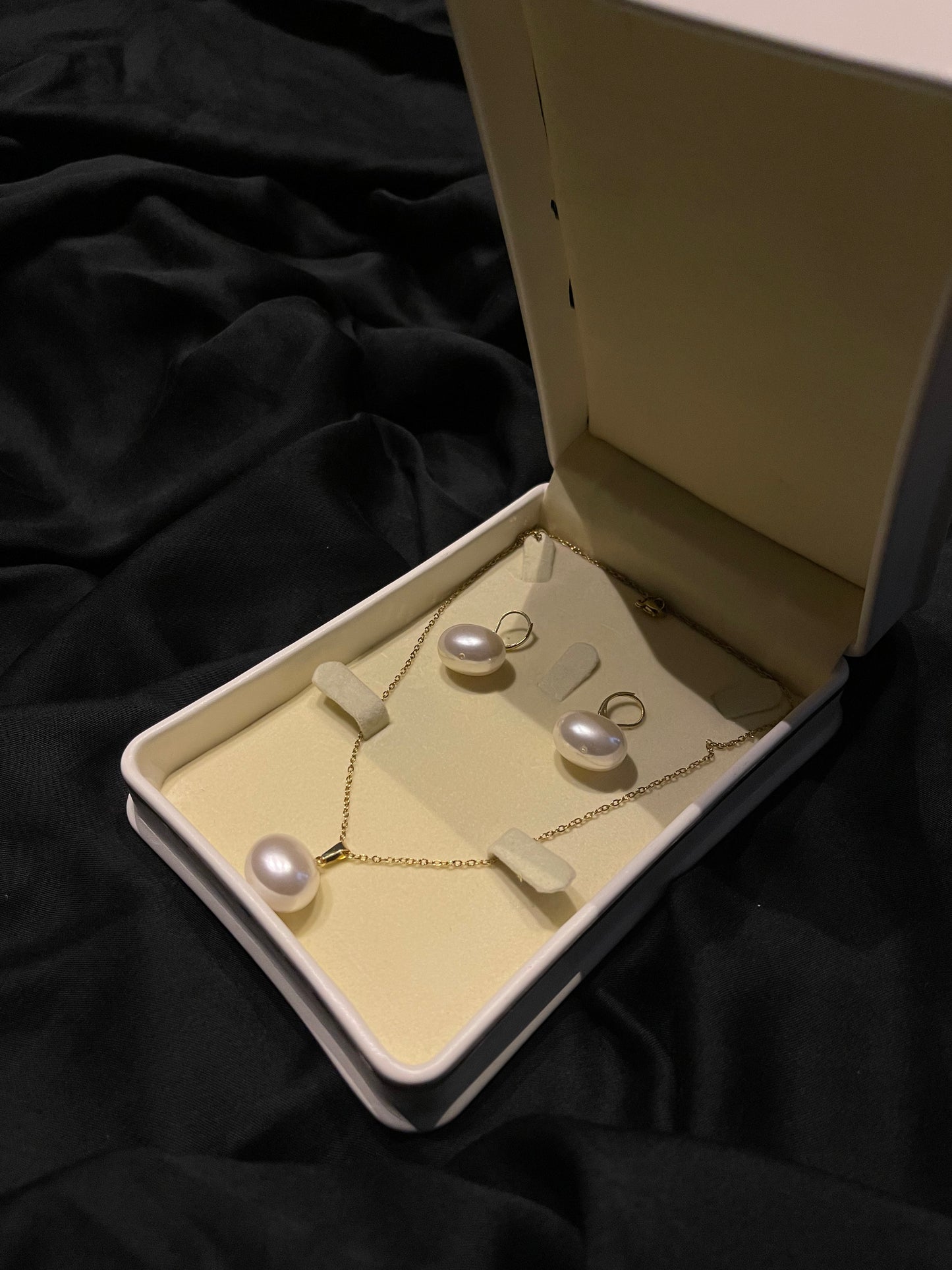 A gold jewelry set with three freshwater pearls, including a necklace and two matching earrings, displayed inside an open cream-colored jewelry box on a black fabric background.