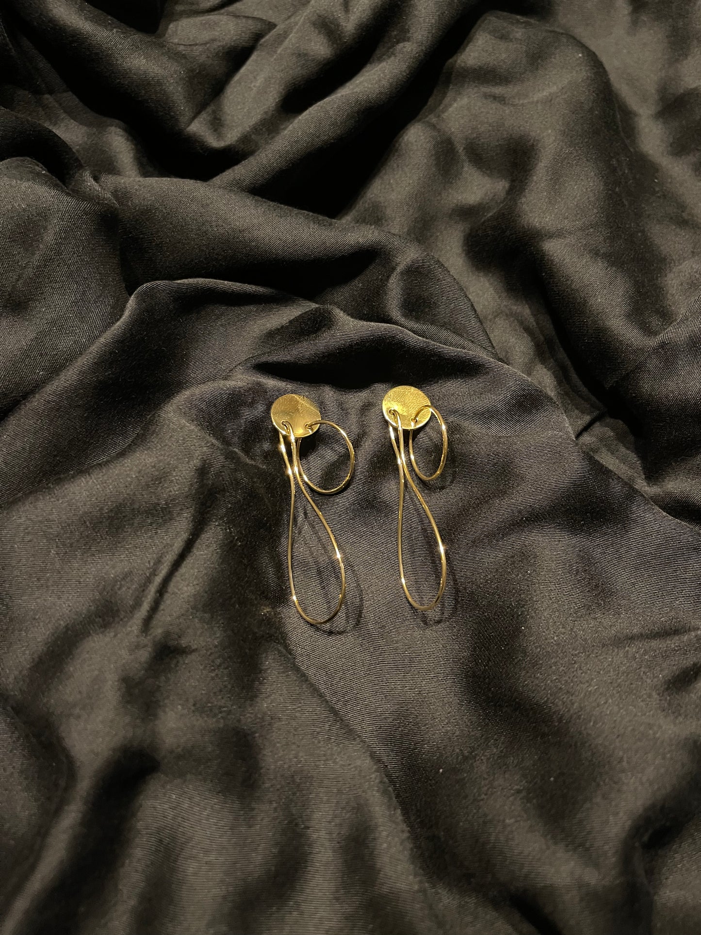 A pair of gold-toned drop earrings with circular disc tops and thin elongated loops, displayed on draped black satin fabric.