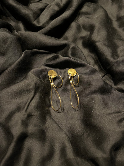 A pair of gold-toned drop earrings with circular disc tops and thin elongated loops, displayed on draped black satin fabric.