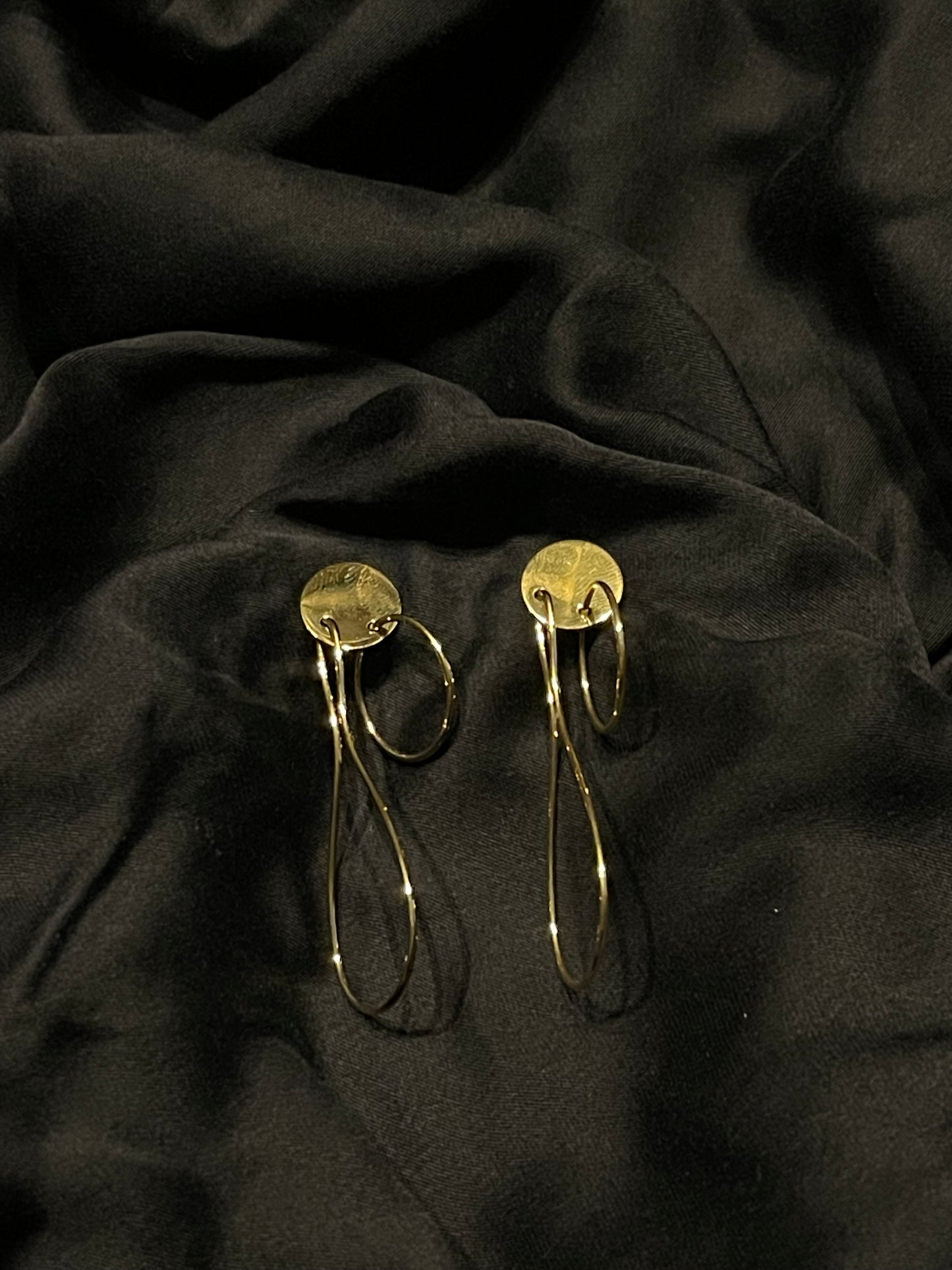 A pair of gold-toned statement earrings with circular disc tops and long, thin loop drops, displayed on draped black fabric.