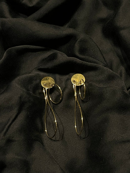 A pair of gold-toned statement earrings with circular disc tops and long, thin loop drops, displayed on draped black fabric.