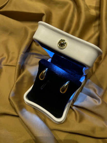 A pair of gold teardrop-shaped drop earrings displayed in an open white jewelry box with a black velvet interior, set against a draped gold satin fabric background.