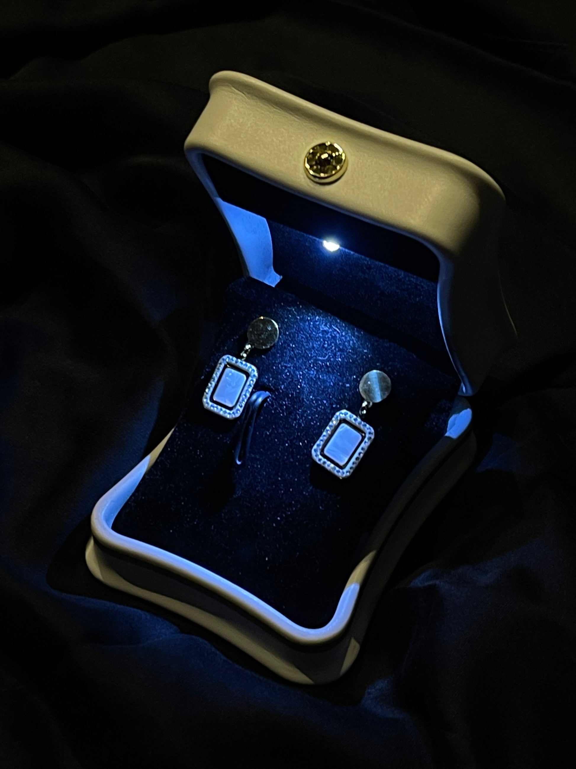 A pair of silver-toned rectangular drop earrings with light blue square inlays and small clear rhinestone borders, displayed inside an open cream jewelry box with a blue interior on a dark fabric background.