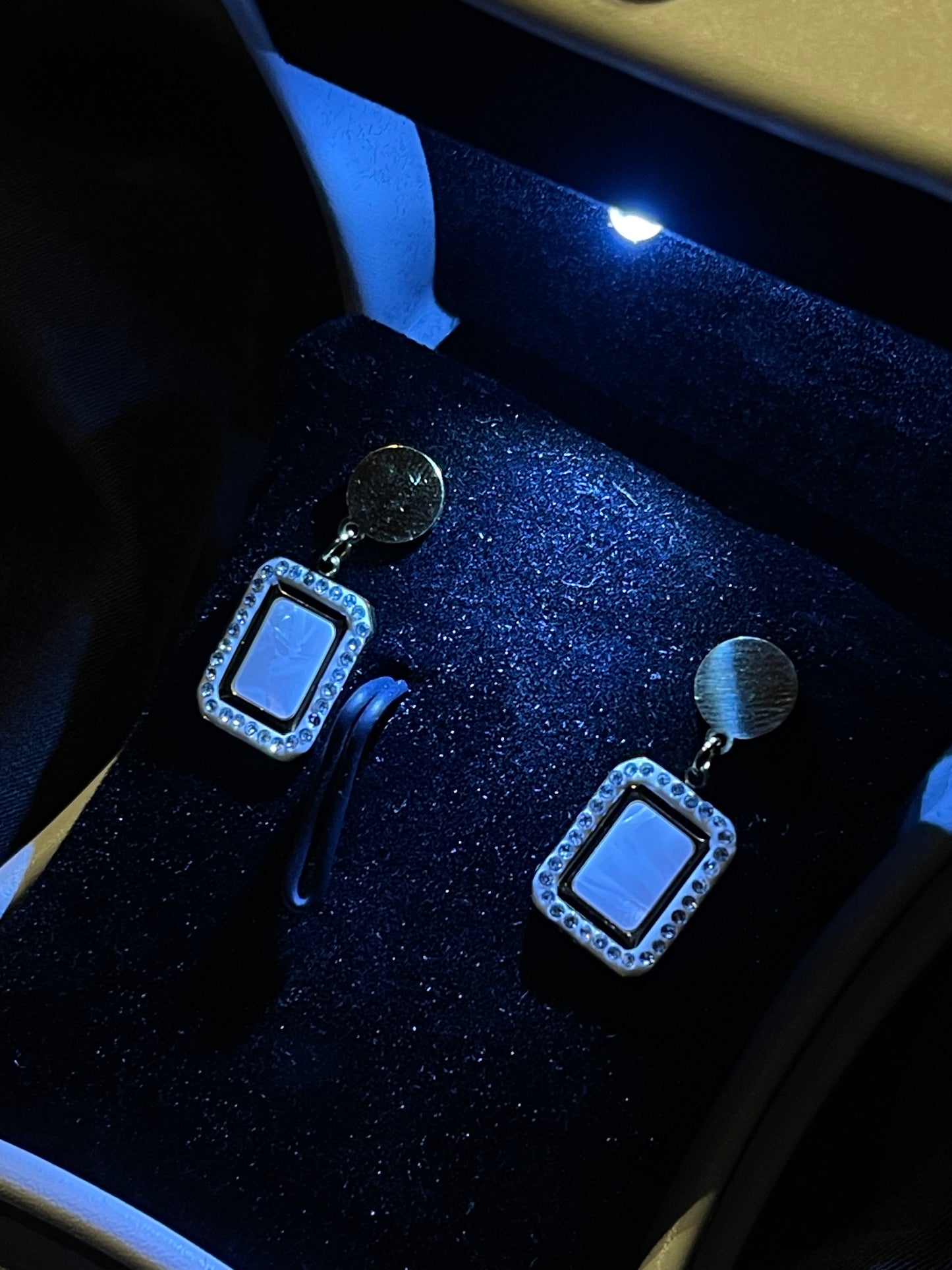 A pair of rectangular drop earrings with silver-toned metal frames and round gold-toned studs, displayed on a black velvet jewelry box under blue light.