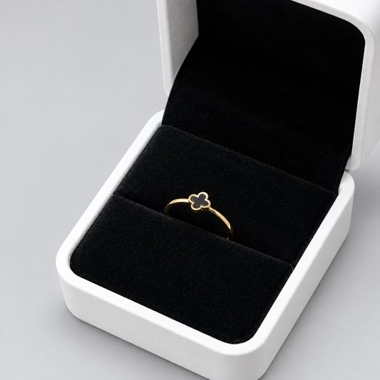 A delicate gold ring with a small clover or flower-like motif, displayed inside an open white jewelry box with a black velvet interior, set against a soft beige fabric background.