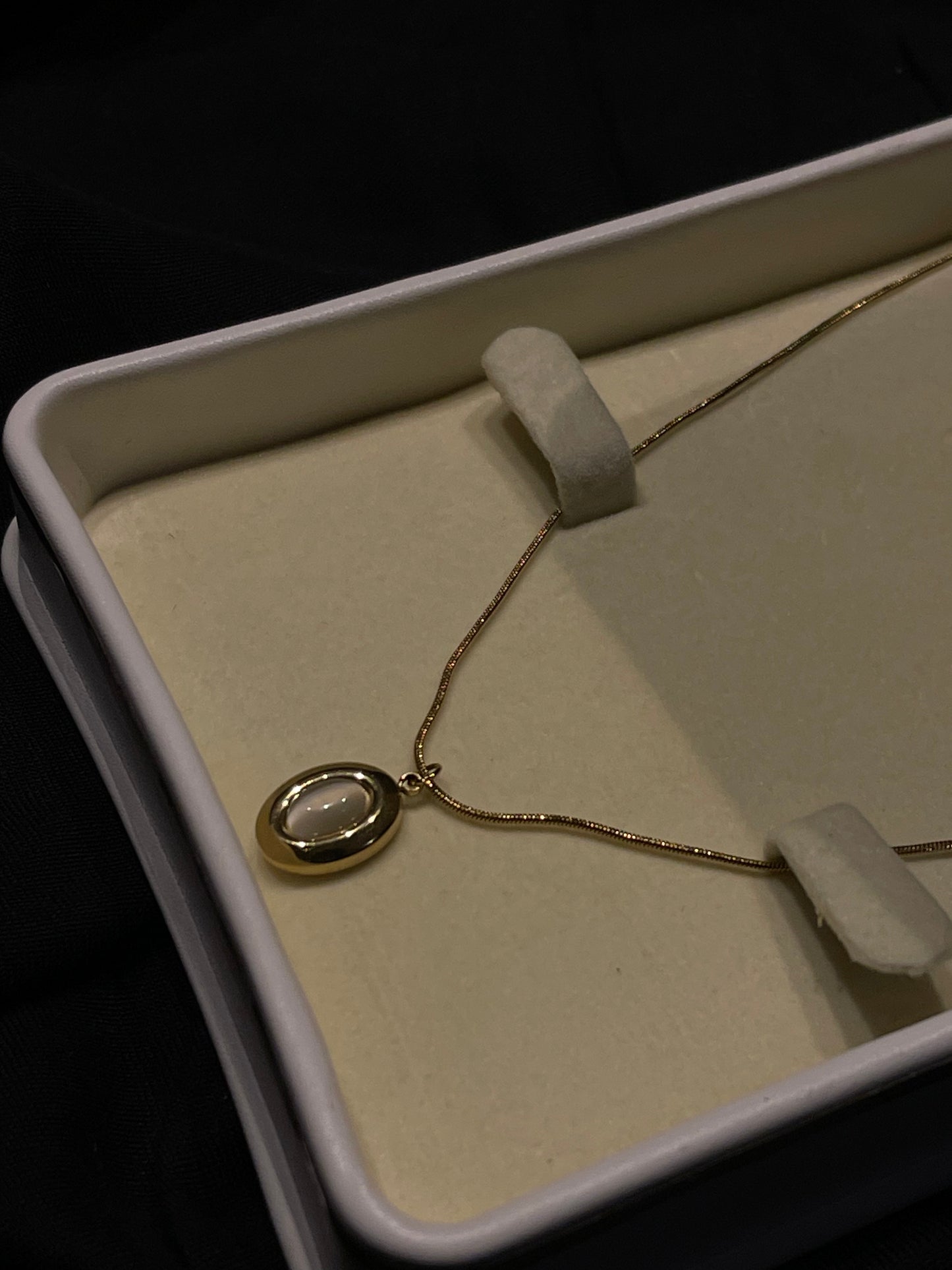 A gold necklace with an oval pendant featuring a white inlay, displayed inside an open cream-colored jewelry box on a dark background.