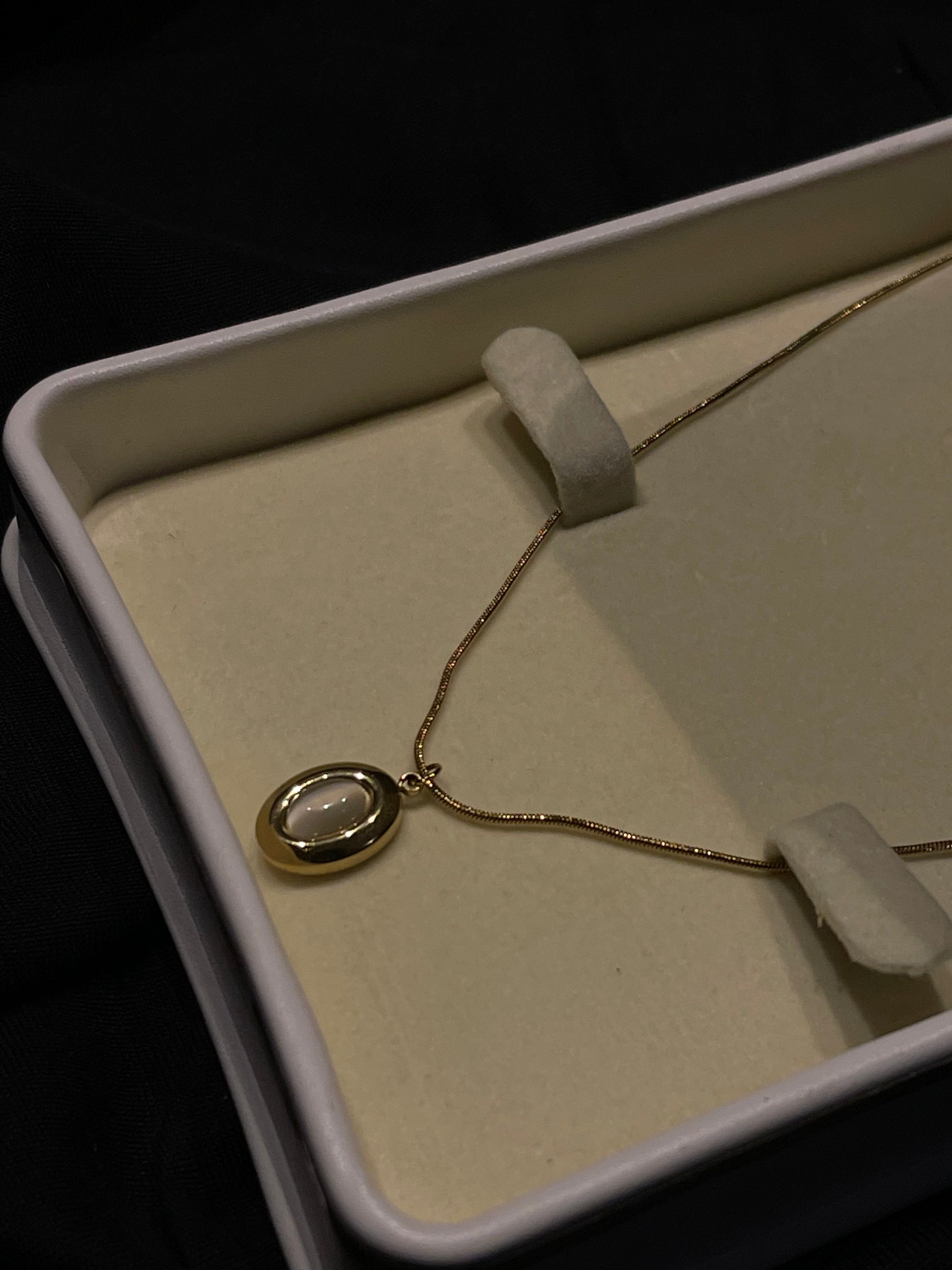 A gold necklace with an oval pendant featuring a white inlay, displayed inside an open cream-colored jewelry box on a dark background.