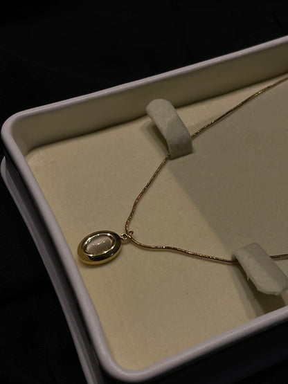 A gold necklace with an oval pendant featuring a white inlay, displayed inside an open cream-colored jewelry box on a dark background.