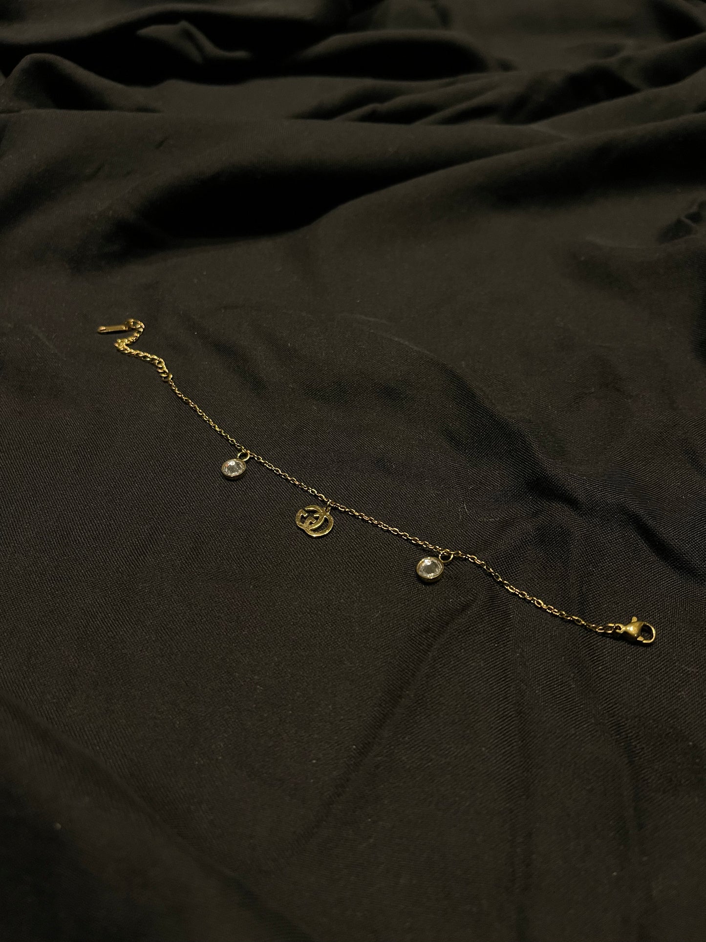 A delicate gold chain anklet with three small hanging charms, including a central circular medallion, displayed on a dark, draped fabric background.