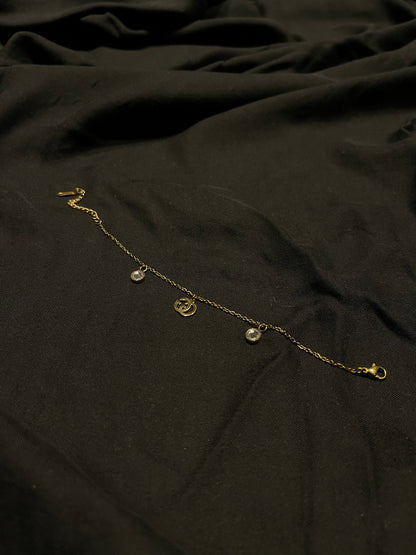 A delicate gold chain anklet with three small hanging charms, including a central circular medallion, displayed on a dark, draped fabric background.