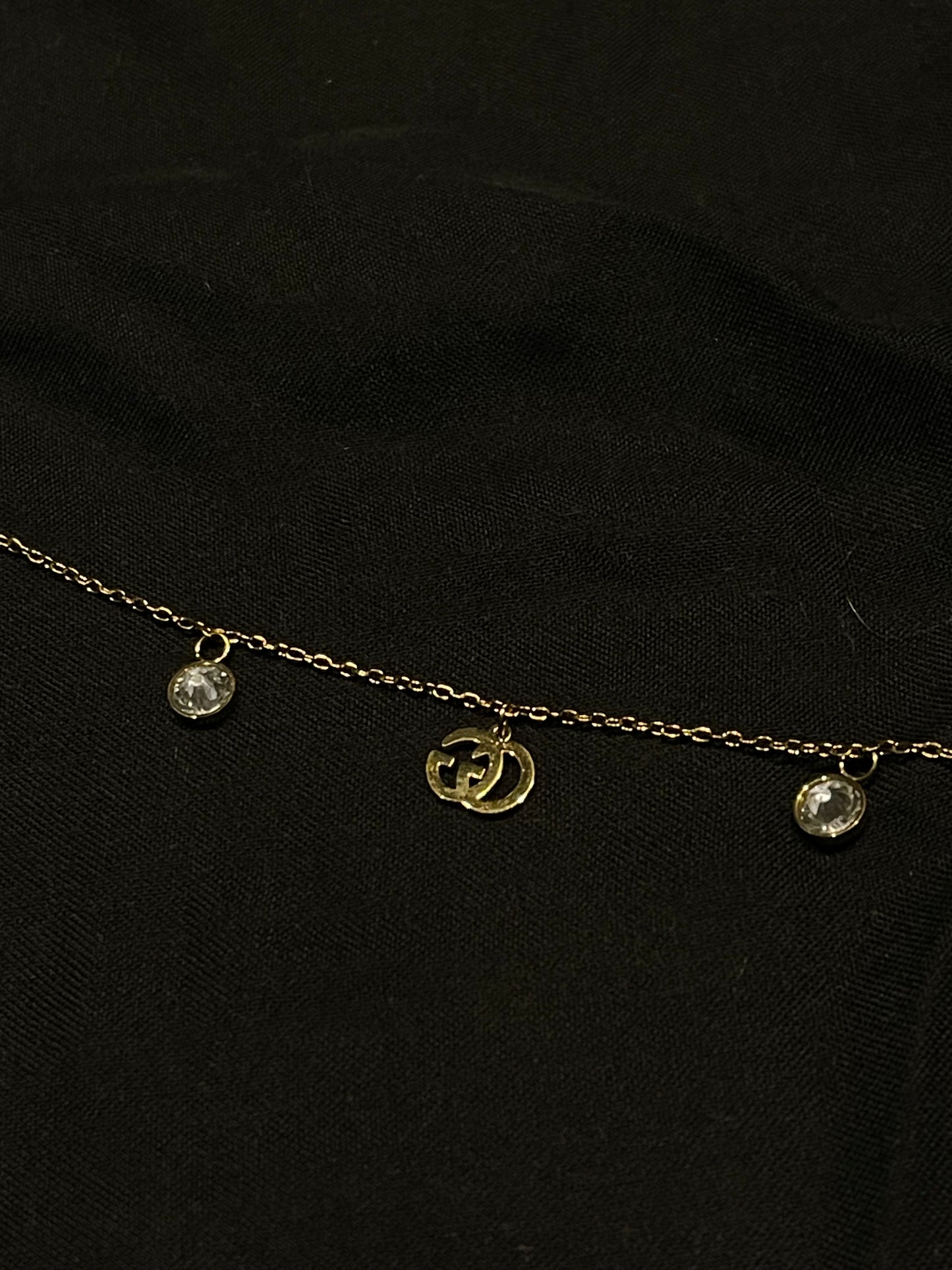 A delicate gold chain necklace featuring a central gold double-G interlocking logo charm and two small clear crystal droplets on either side, displayed on a black fabric background.