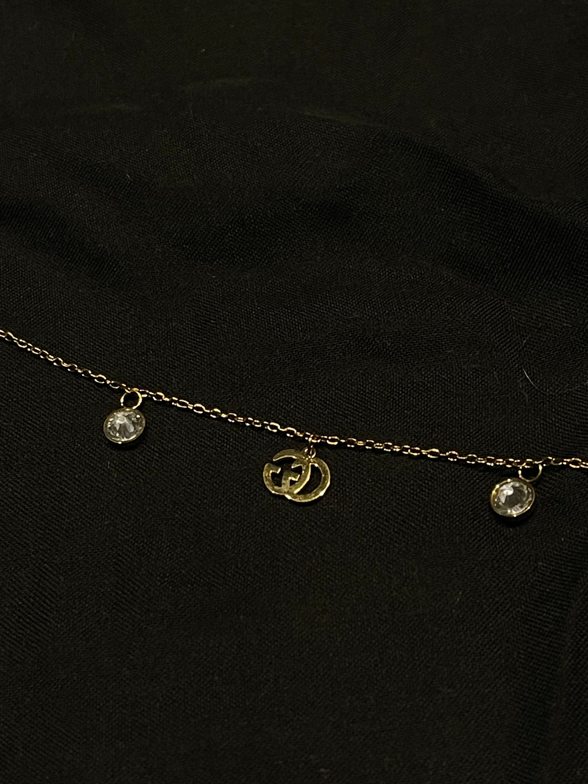A delicate gold chain necklace featuring a central gold double-G interlocking logo charm and two small clear crystal droplets on either side, displayed on a black fabric background.