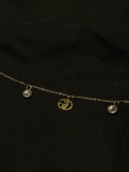 A delicate gold chain necklace featuring a central gold double-G interlocking logo charm and two small clear crystal droplets on either side, displayed on a black fabric background.