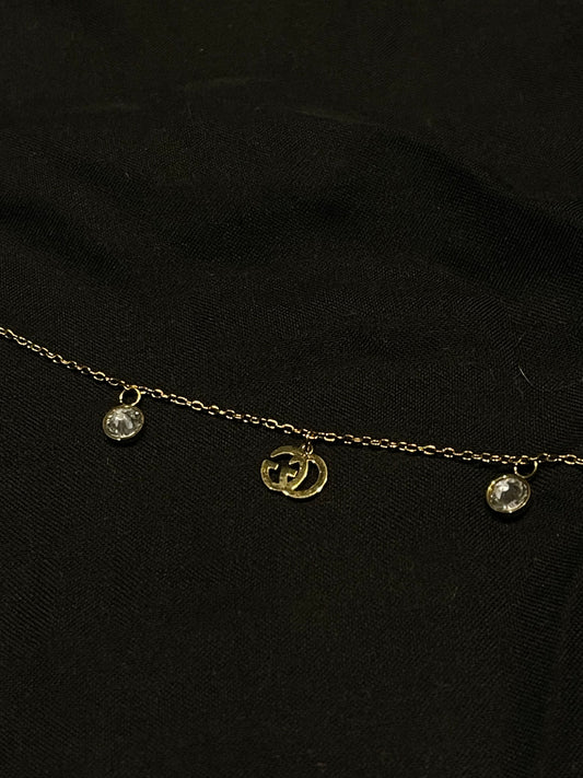 A delicate gold chain necklace featuring a central gold double-G interlocking logo charm and two small clear crystal droplets on either side, displayed on a black fabric background.