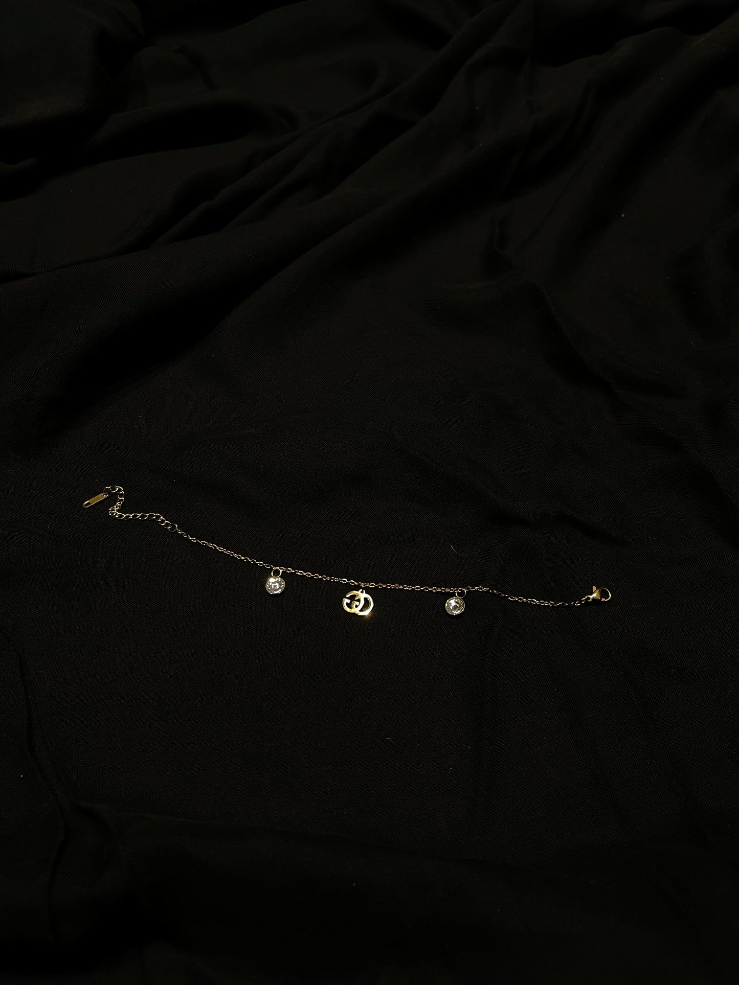 A delicate gold chain anklet with three small dangling charms, including a central crescent moon and two round beads, laid flat on a black fabric background.