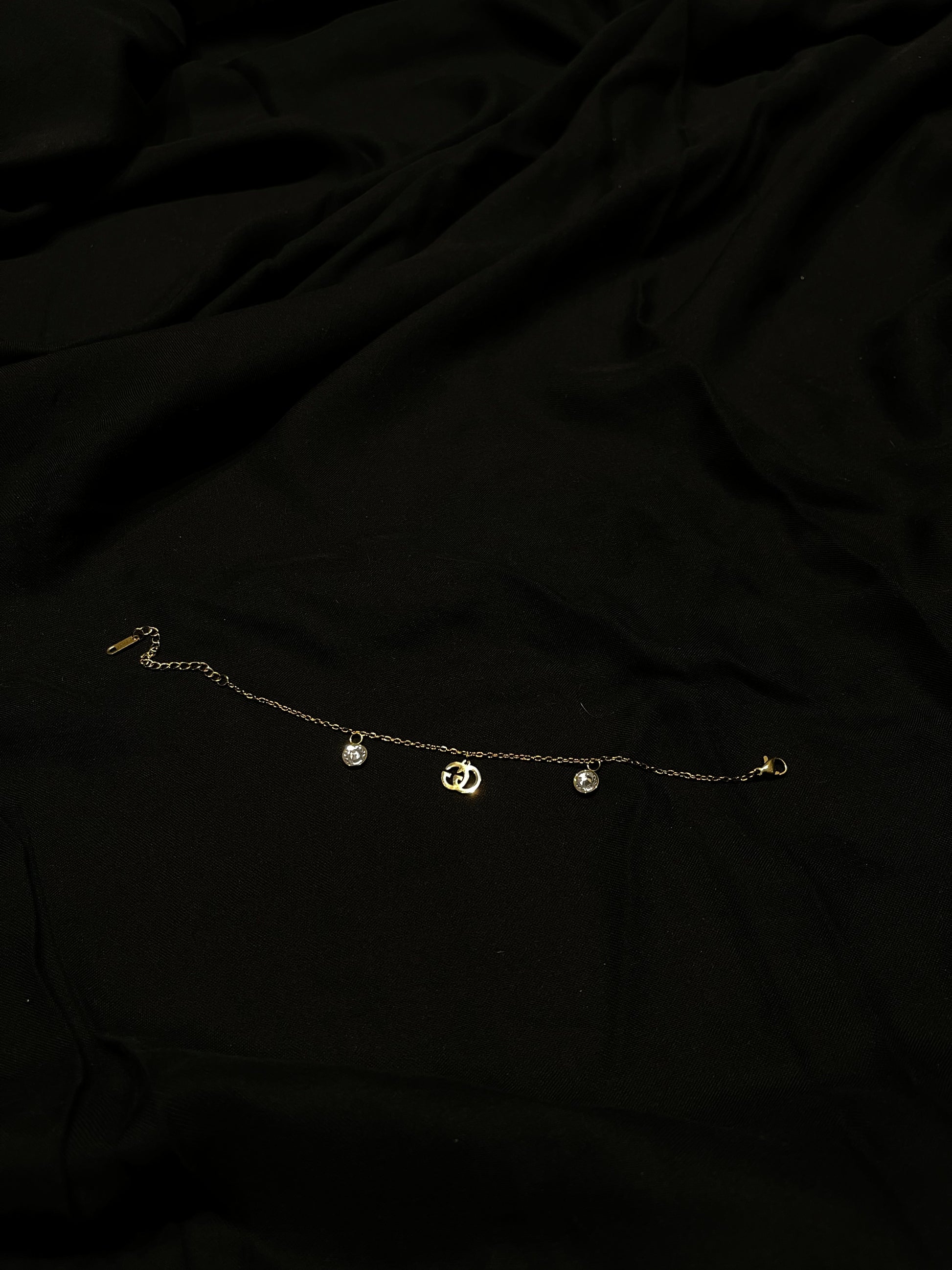 A delicate gold chain anklet with three small dangling charms, including a central crescent moon and two round beads, laid flat on a black fabric background.