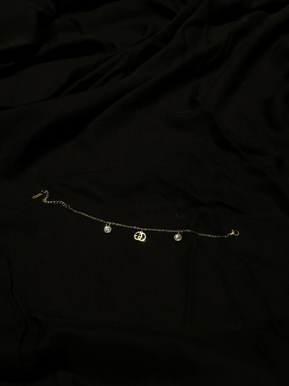 A delicate gold chain anklet with three small dangling charms, including a central crescent moon and two round beads, laid flat on a black fabric background.