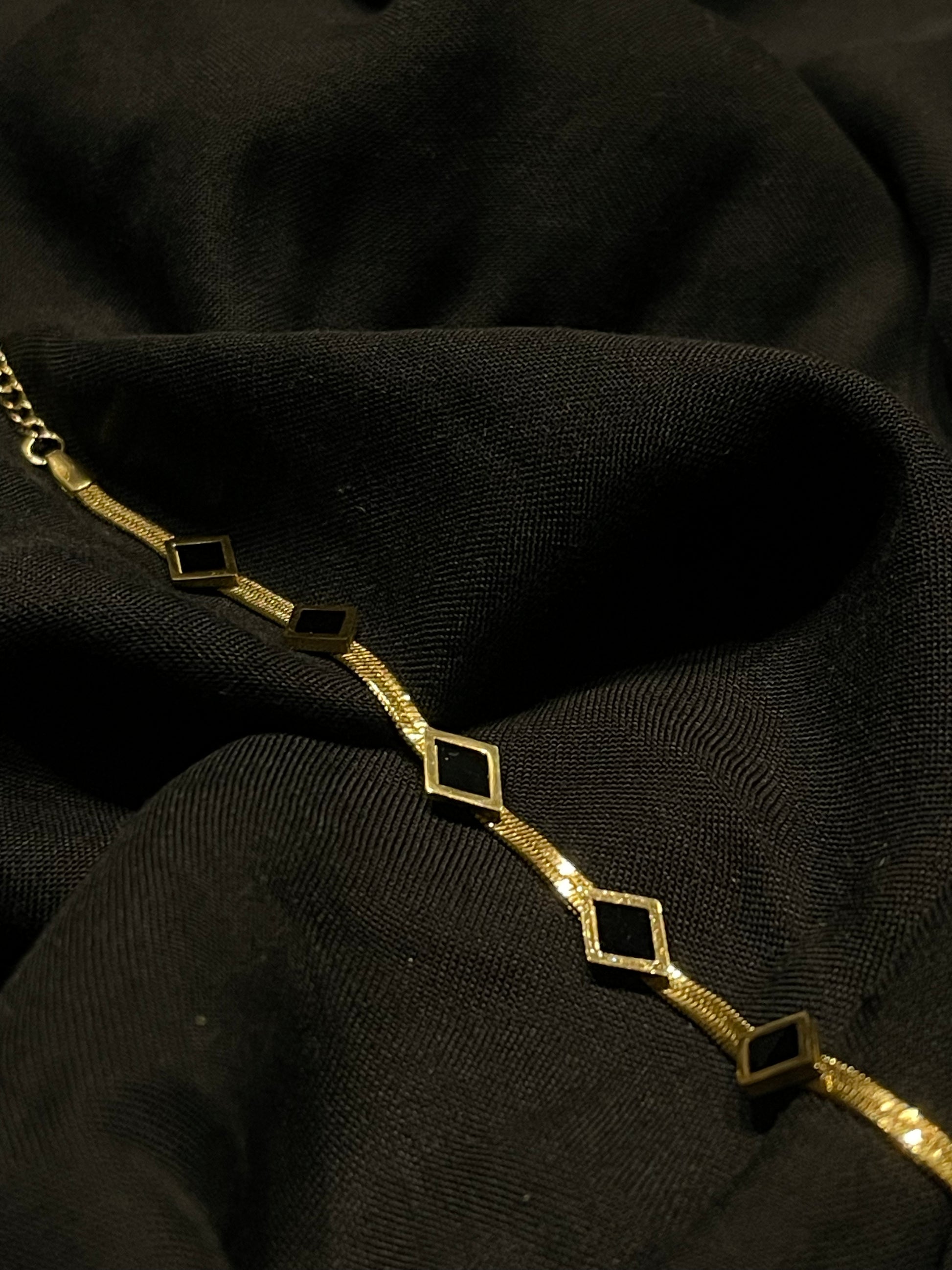 A gold chain bracelet featuring repeating open diamond-shaped links, displayed on a textured black fabric background.
