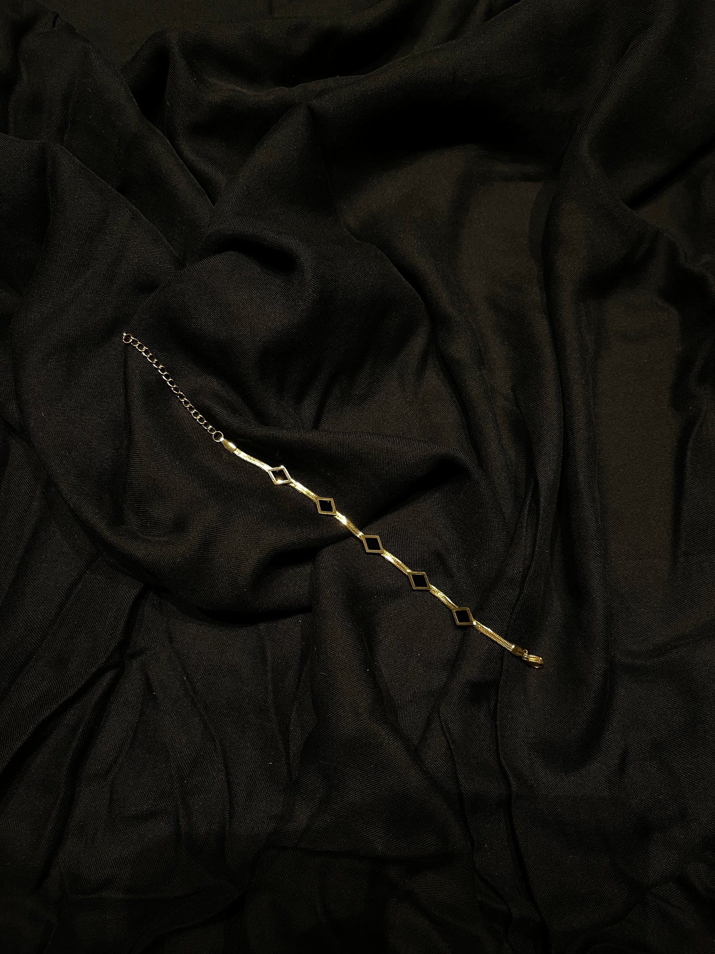 A delicate gold chain bracelet with repeating open diamond-shaped links, laid diagonally across draped black fabric.