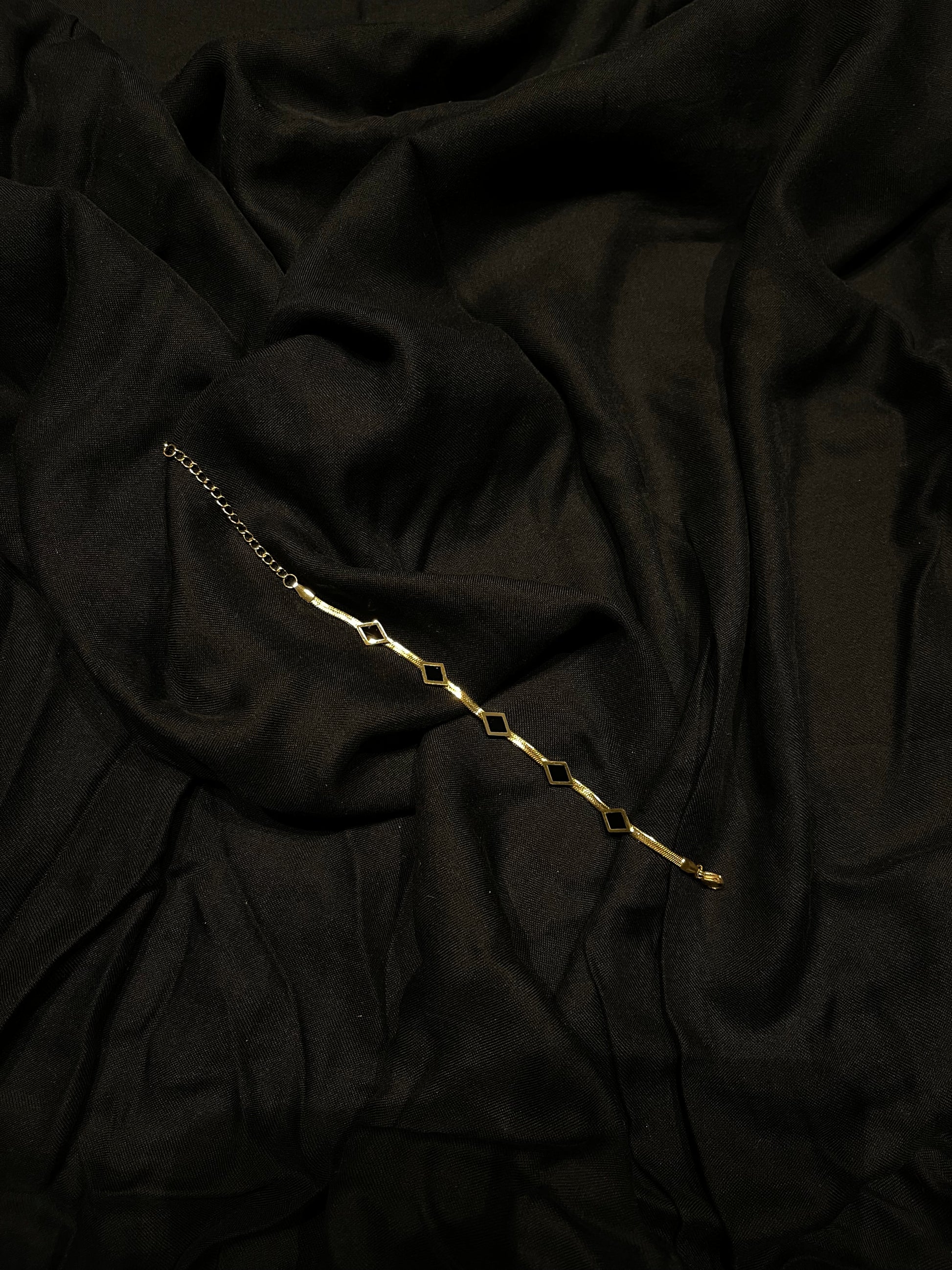 A delicate gold chain bracelet with repeating open diamond-shaped links, laid diagonally across draped black fabric.
