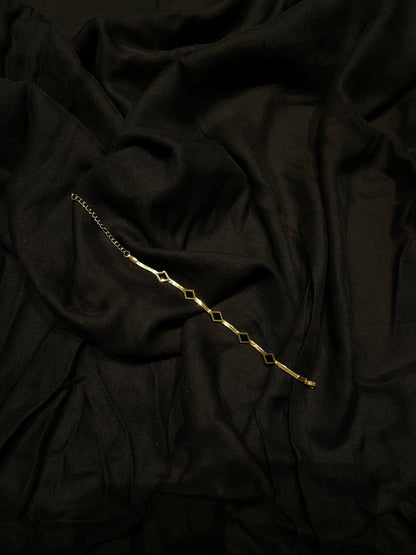 A delicate gold chain bracelet with repeating open diamond-shaped links, laid diagonally across draped black fabric.