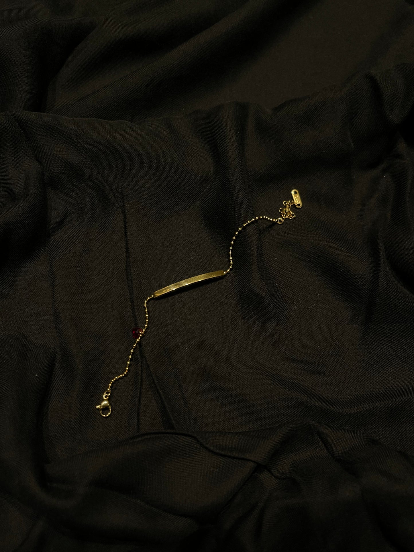 A gold-toned chain bracelet with a flat rectangular bar pendant, laid on draped black fabric.