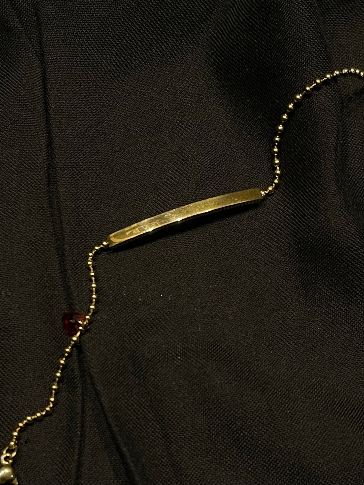 A gold bar necklace with a matching gold beaded chain, laid out on a textured black fabric background.
