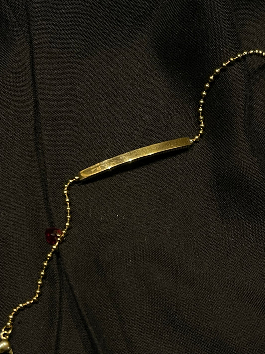 A gold bar necklace with a matching gold beaded chain, laid out on a textured black fabric background.