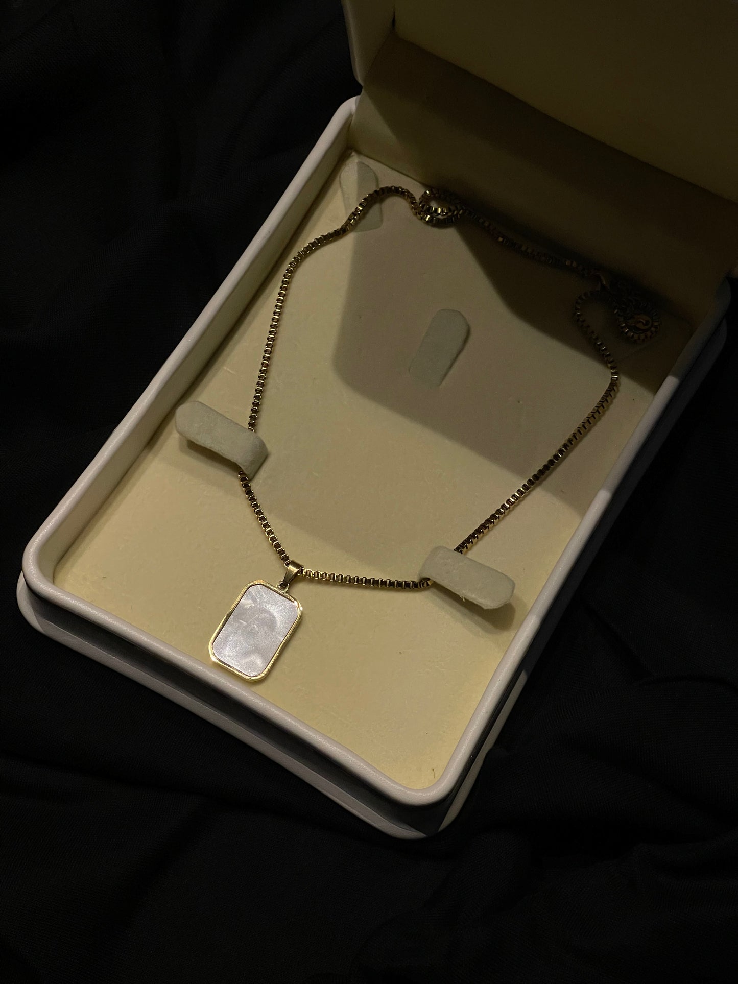 A gold-tone chain necklace with a rectangular pendant made of iridescent mother-of-pearl, presented inside an open cream-colored jewelry box on a black background.