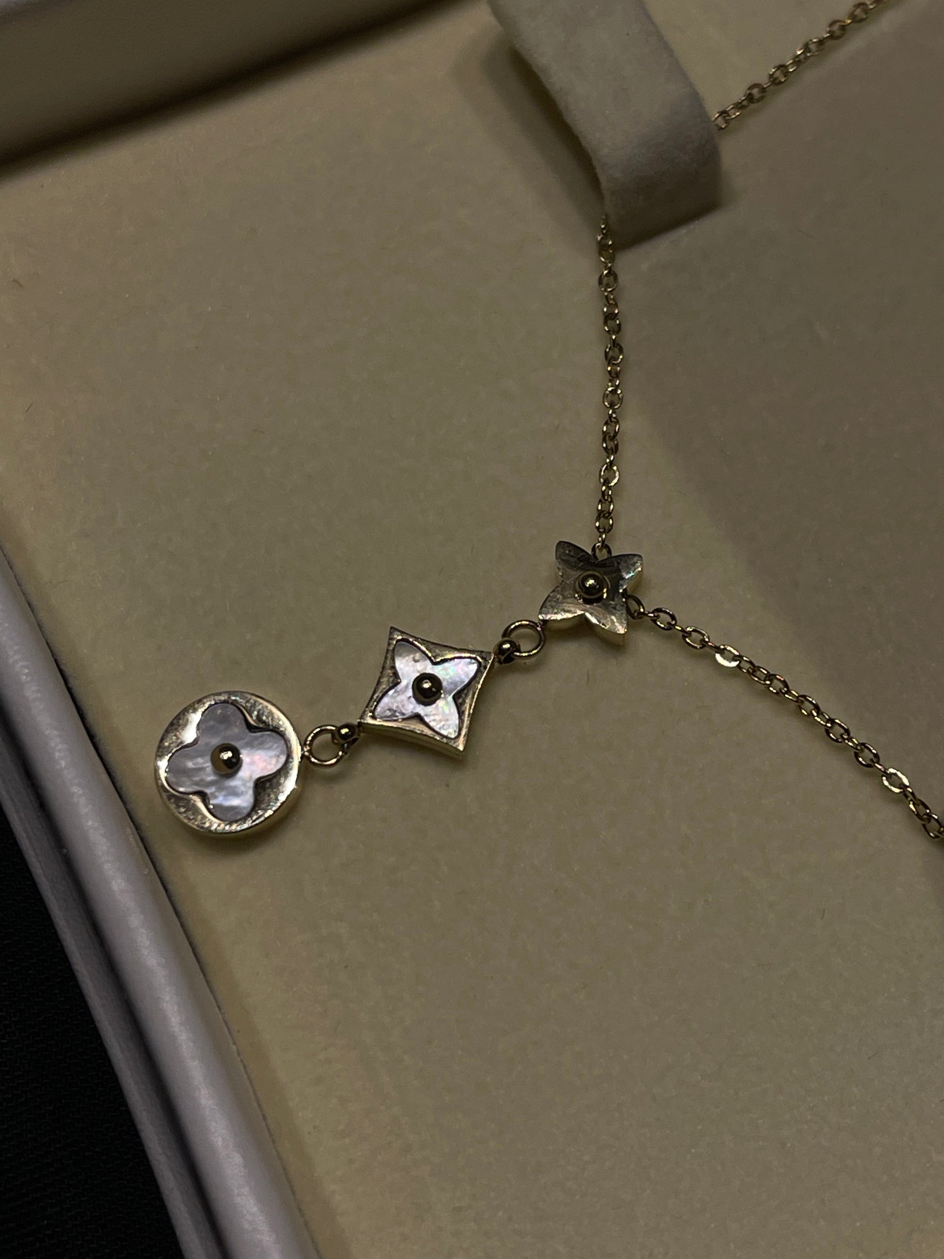 A silver-toned necklace with three interlocking floral charms, featuring one round charm with a white mother-of-pearl flower center, one diamond-shaped charm with a matching center, and one smaller butterfly-like charm. The necklace is displayed inside a beige jewelry box.