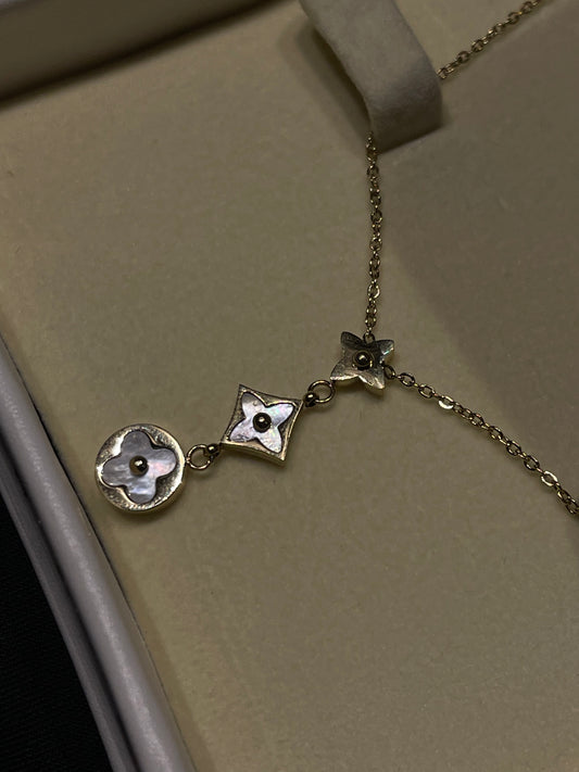A silver-toned necklace with three interlocking floral charms, featuring one round charm with a white mother-of-pearl flower center, one diamond-shaped charm with a matching center, and one smaller butterfly-like charm. The necklace is displayed inside a beige jewelry box.