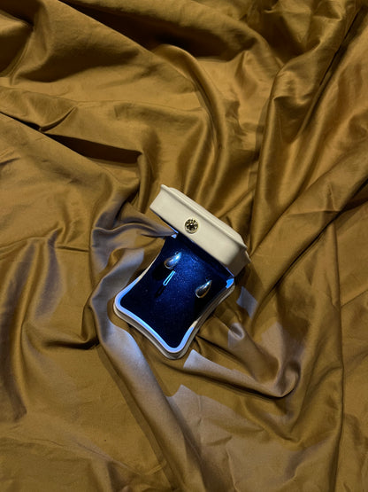 A pair of silver hoop earrings displayed in an open white jewelry box with a blue velvet interior, placed on draped gold satin fabric.