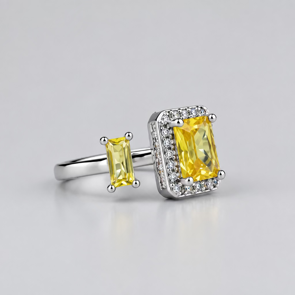 A silver ring featuring a large rectangular yellow gemstone and a smaller matching yellow stone, displayed on a light surface with a blurred white jewelry box in the background.