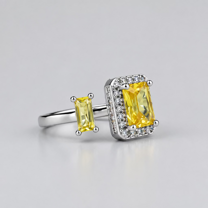 A silver ring featuring a large rectangular yellow gemstone and a smaller matching yellow stone, displayed on a light surface with a blurred white jewelry box in the background.