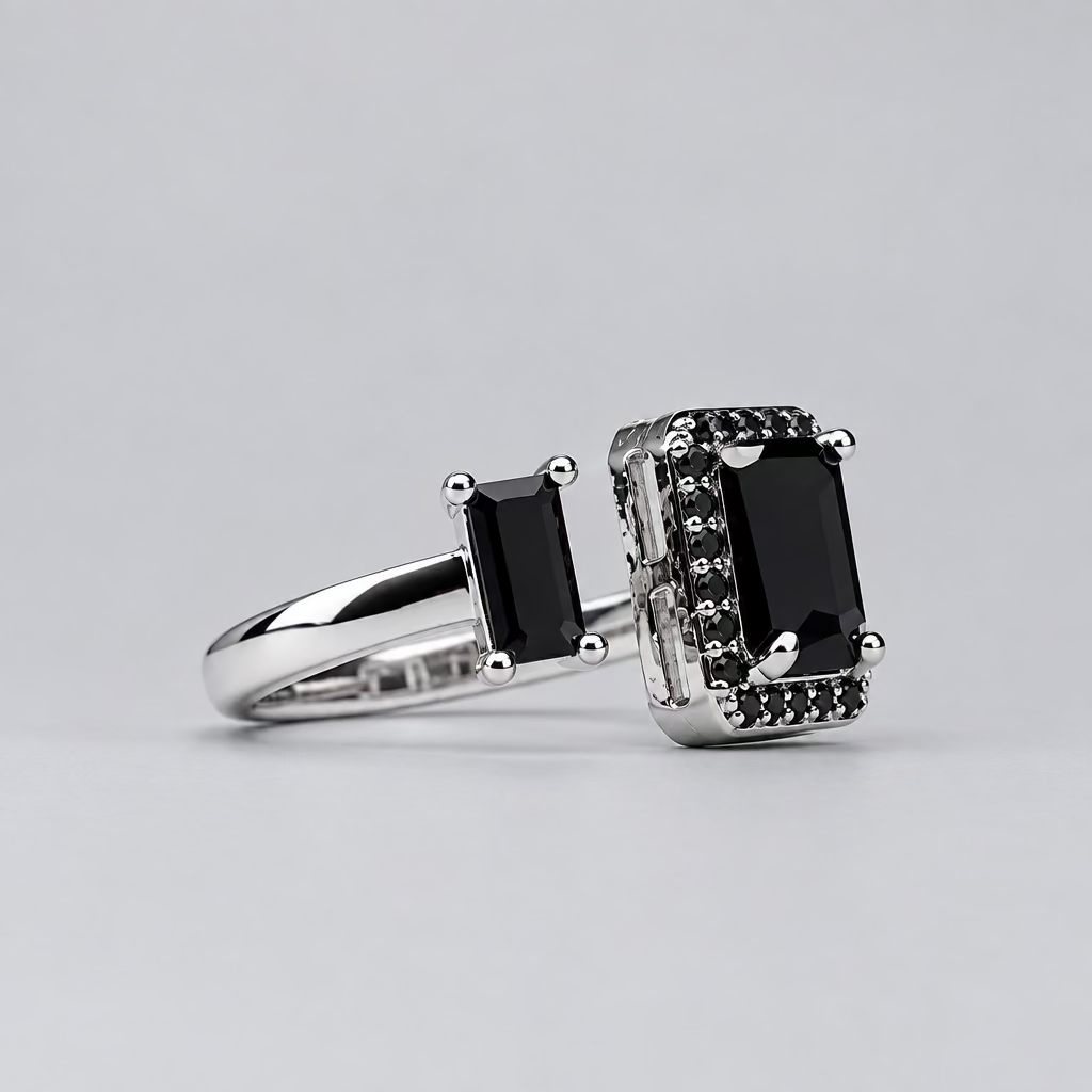 A silver-toned ring with a large black rectangular center stone and smaller black stones on the shoulders, displayed in front of a blurred white jewelry box with an orange logo.