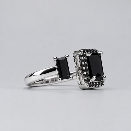 A silver-toned ring with a large black rectangular center stone and smaller black stones on the shoulders, displayed in front of a blurred white jewelry box with an orange logo.
