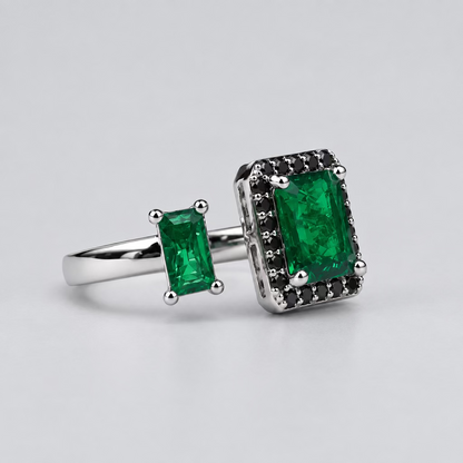 A silver ring featuring a large rectangular green emerald-cut center stone and a smaller matching green stone on the band, displayed in front of a blurred white jewelry box with an orange logo.