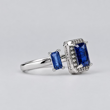 A silver ring with rectangular blue gemstones, displayed on a light surface with a blurred white jewelry box in the background featuring the 'Meenu' brand logo.