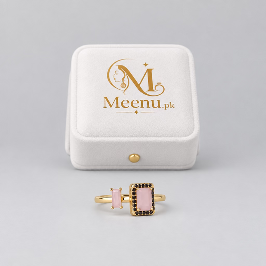 A rose gold ring with a rectangular pink stone and black accent stones, displayed in front of its white Meenu Jewels box on a light purple surface.